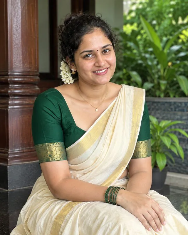Aarti in a cream and gold kasavu saree with a green silk blouse, jasmine in her hair, seated on a wooden veranda